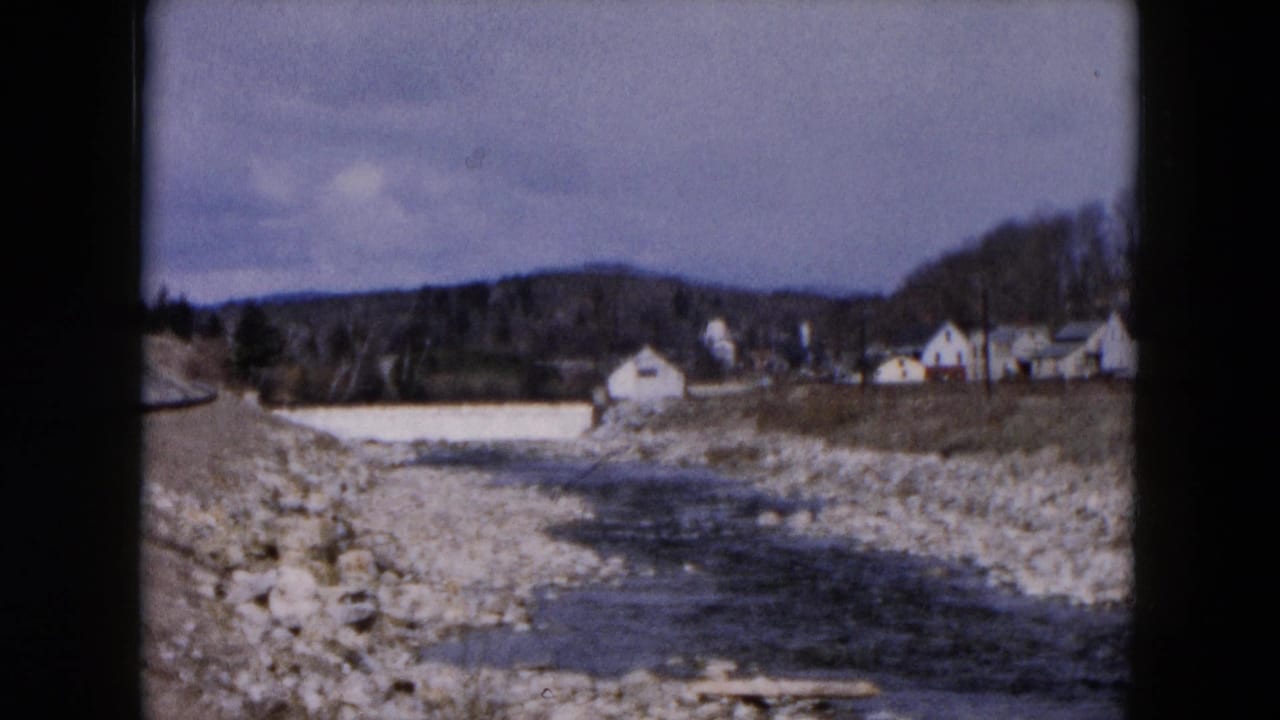 Stream and gravel riverbed curve past a road edge, with houses and wooded hills beyond.
