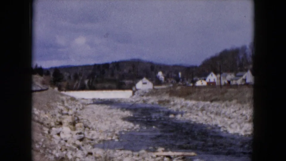 1957 Hicksville roadside landscape with a rocky streambed, water channel, distant white buildings, trees, and hills.