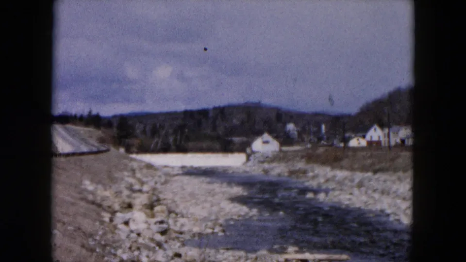 1957 8mm roadside view in Hicksville showing a dirt road, guardrail, rocky streambed, distant buildings, and wooded hills.