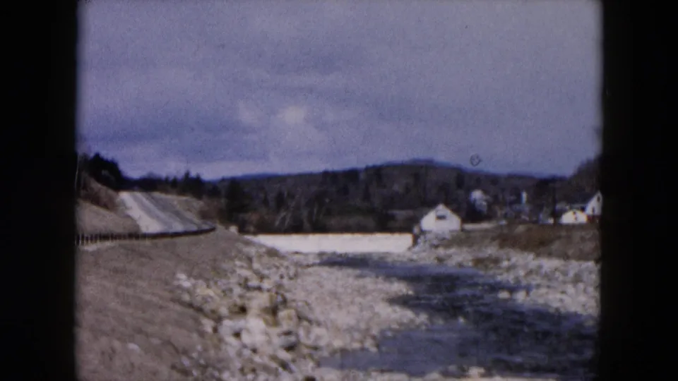 1957 Hicksville landscape with a guardrail along the road and a rocky streambed leading toward distant buildings and hills.