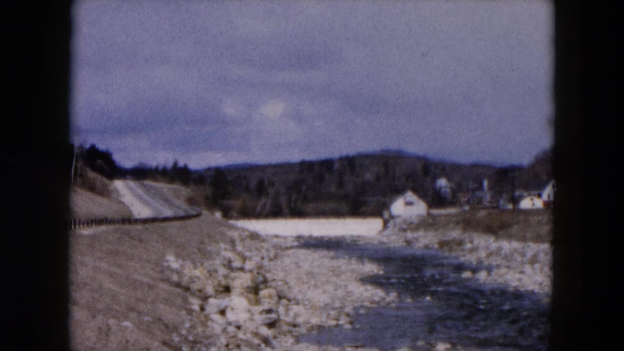 Rocky stream beside a rural road with distant houses under an overcast sky.