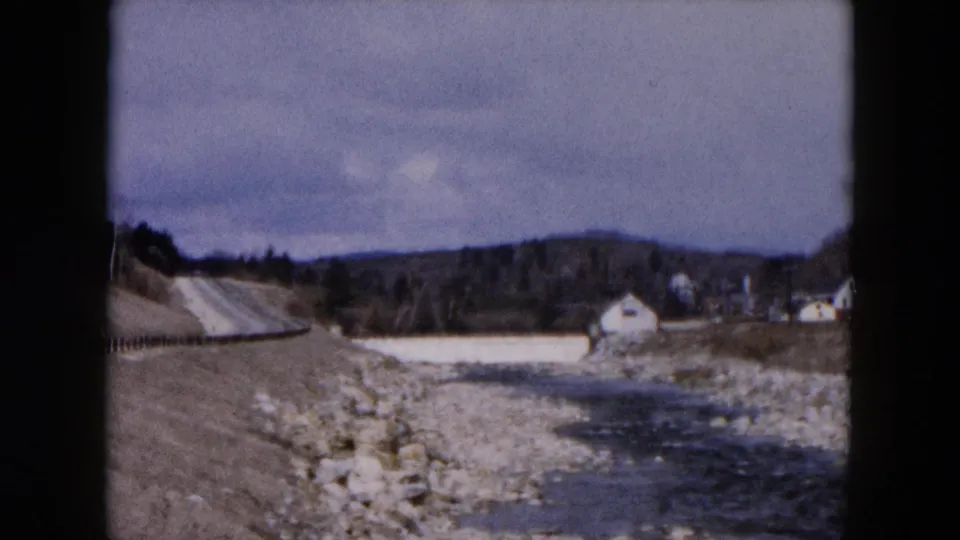 1957 Hicksville home-movie frame showing a rocky stream, roadside edge, distant houses, low hills, and cloudy sky.