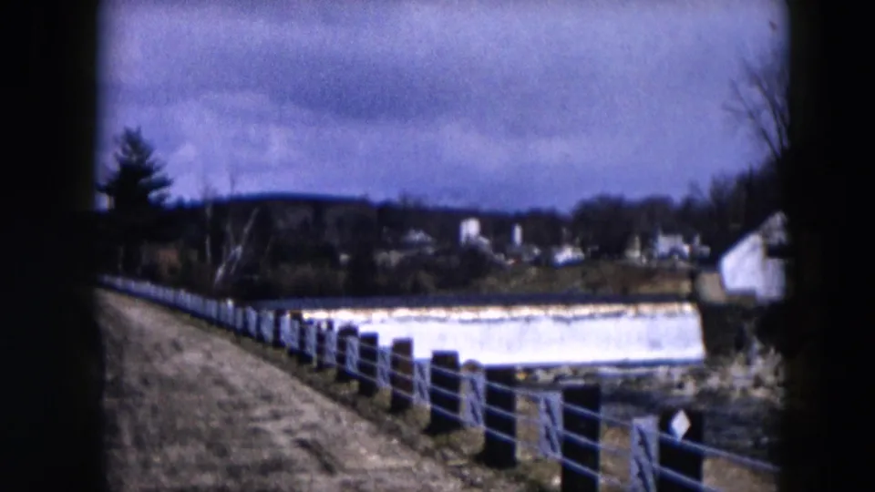 1957 Hicksville frame showing a white dam or barrier along a stream, fence posts, a road edge, distant houses, and cloudy sky.