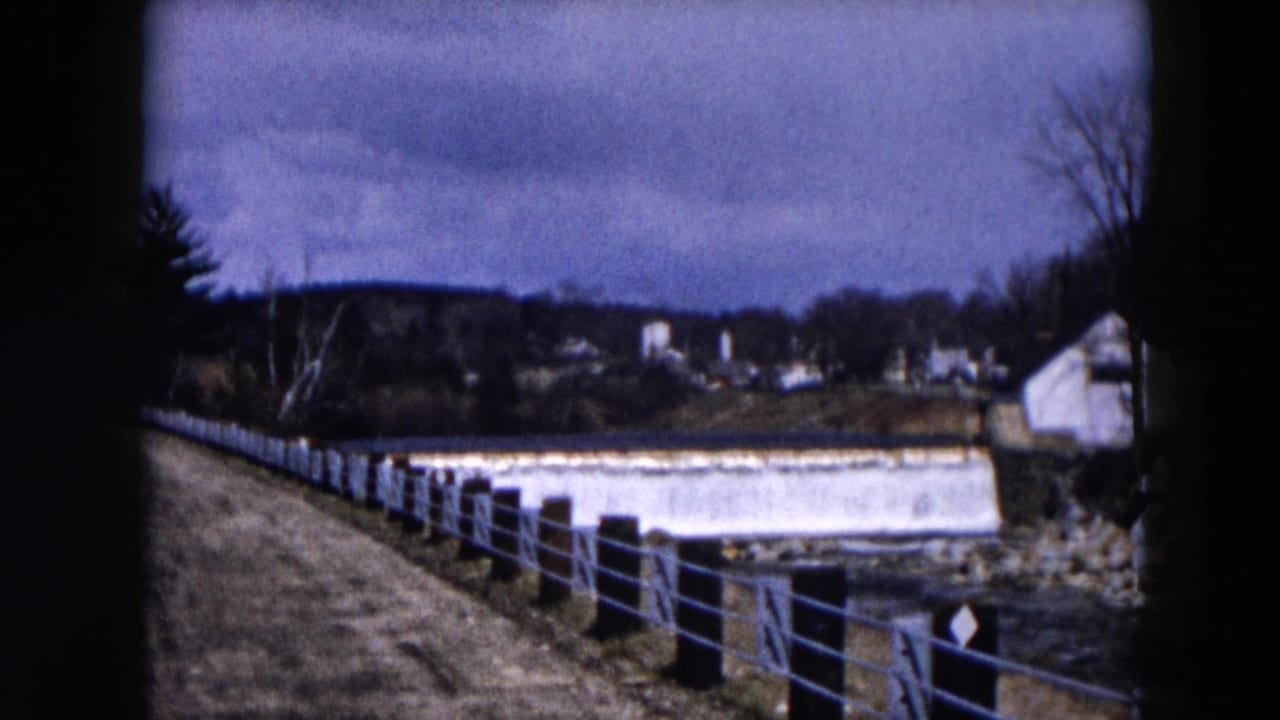 1957 Hicksville, New York 8mm frame showing a concrete river dam, metal railing, bare trees, and distant buildings.
