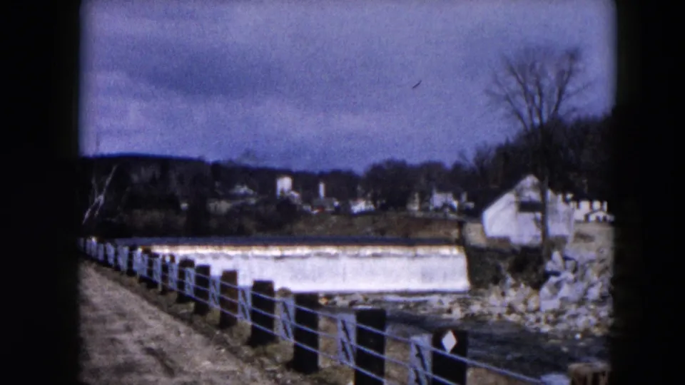 1957 Hicksville 8mm view of water crossing a low dam with a small riverside building and bare trees beyond.