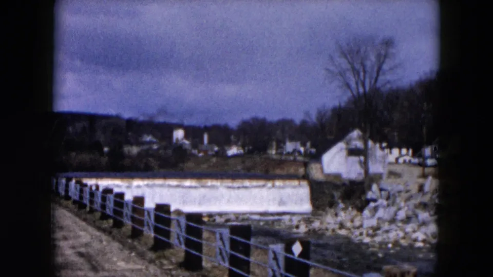 Home-movie frame from Hicksville, New York showing the dam and fenced river path under a cloudy sky in 1957.