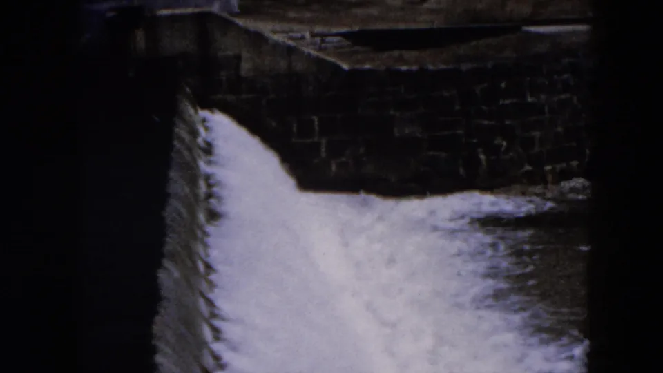 1957 Hicksville 8mm frame showing turbulent water running past dark stonework in a man-made channel.