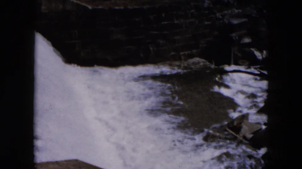 1957 Hicksville home-movie frame with foaming water, dark masonry, and scattered rocks near the channel edge.