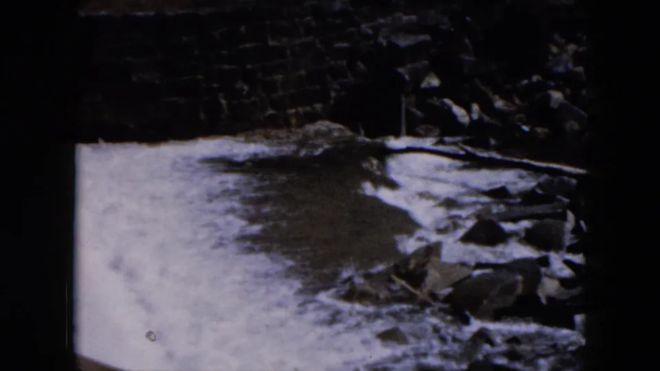 1957 Hicksville 8mm frame showing a wider section of water moving through a stone-lined spillway with rocks nearby.