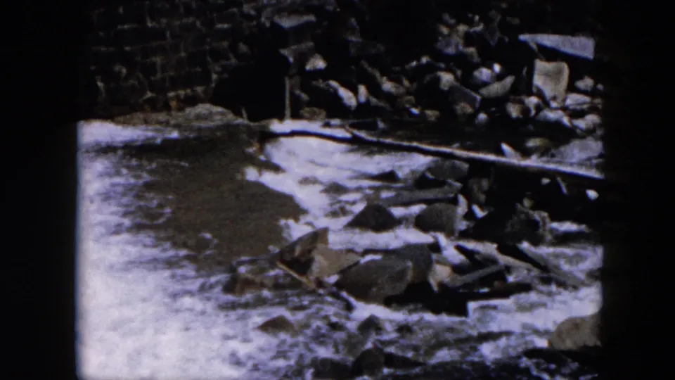 1957 Hicksville frame with turbulent water, a low stone edge, and rubble along the side of the channel.