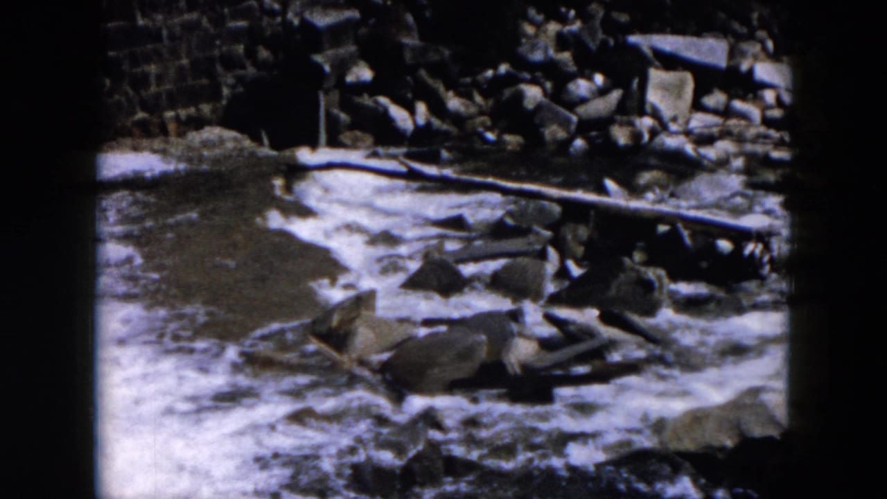 1957 8mm home-movie view of rushing water moving around rocks, with a rough stone edge or wall behind the stream.