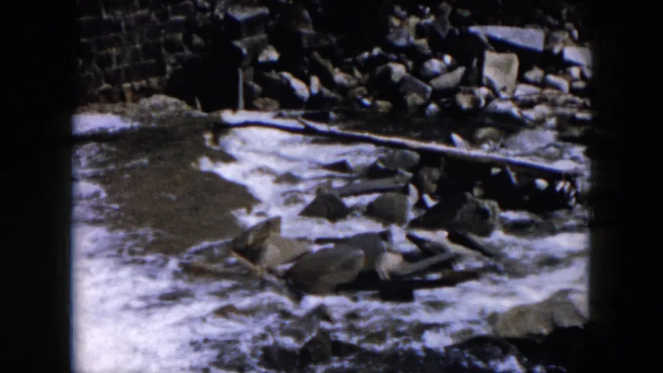 1957 8mm home-movie view of rushing water moving around rocks, with a rough stone edge or wall behind the stream.