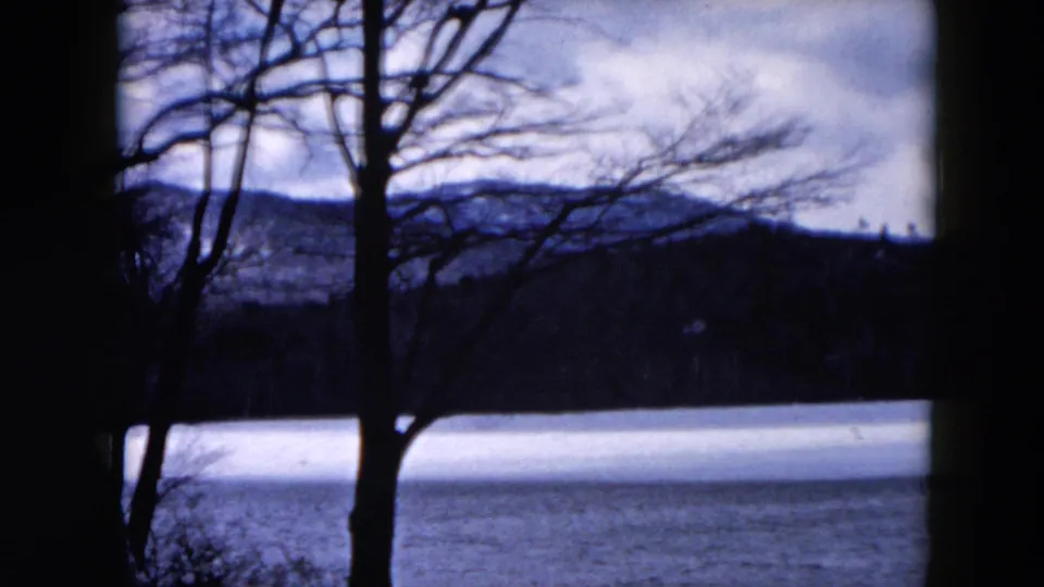 Handheld 1957 8mm view of bare lakeside trees, a calm water surface, wooded hills, and a cloudy sky.