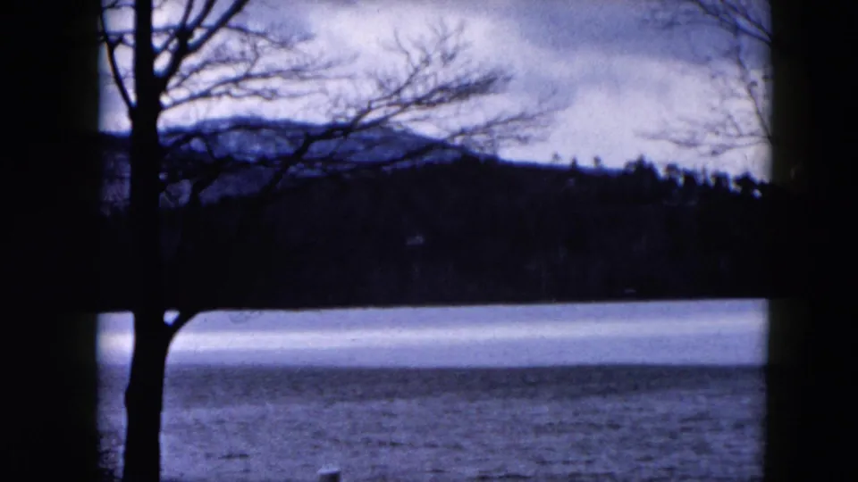 1957 Hicksville, New York 8mm frame showing bare trees, a cold lake surface, distant woods, and low hills under cloudy sky.