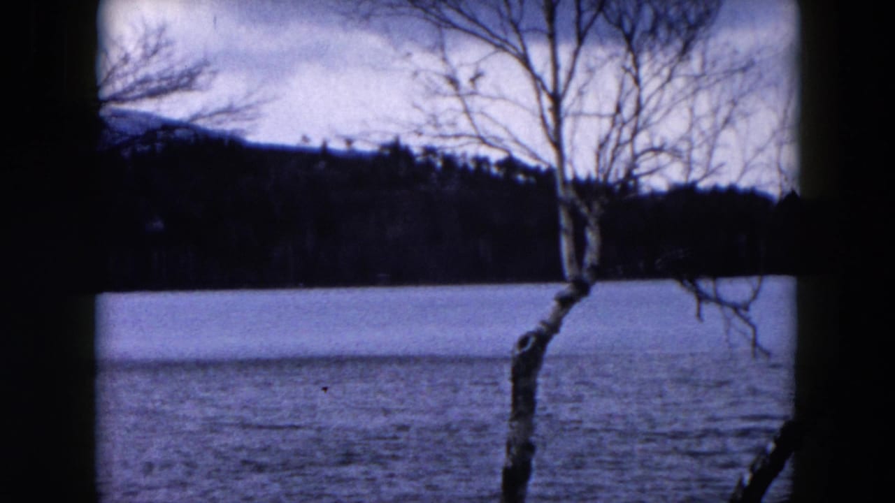 1957 Hicksville, New York home-movie frame showing a cold lake surface, bare trees, distant woods, and low hills.