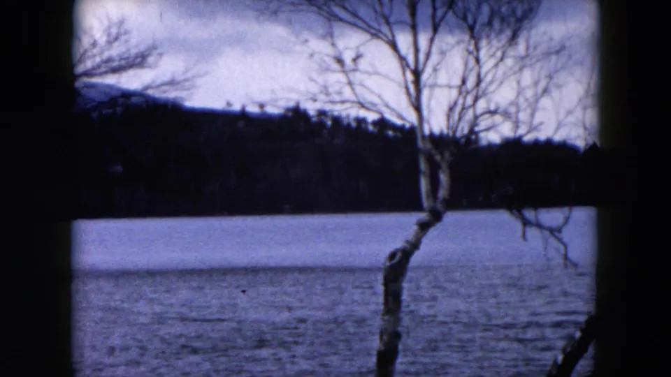 1957 Hicksville, New York home-movie frame showing a cold lake surface, bare trees, distant woods, and low hills.