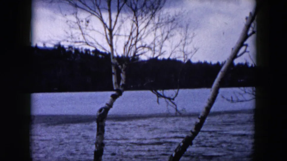 1957 Hicksville, New York frame of a partially frozen lake, leafless trees, and a wooded shoreline in blue-toned 8mm color.