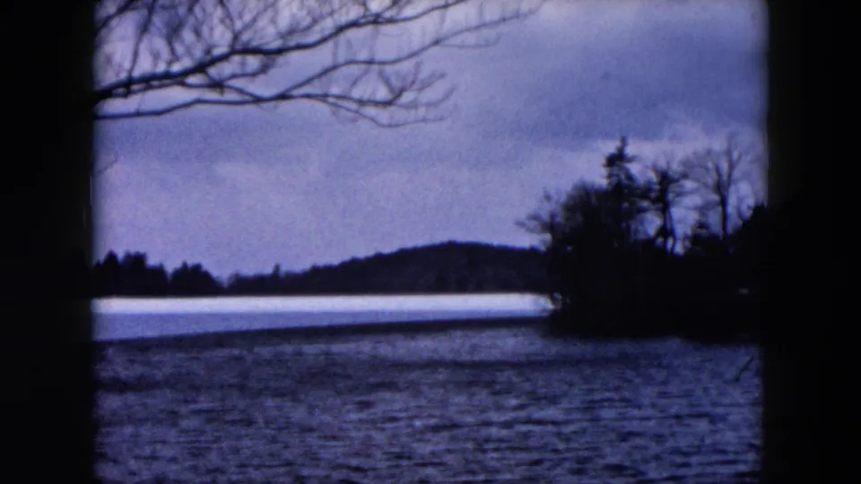 1957 Hicksville, New York home-movie frame showing a lake, distant hills, bare branches, and a small wooded point along the shore.