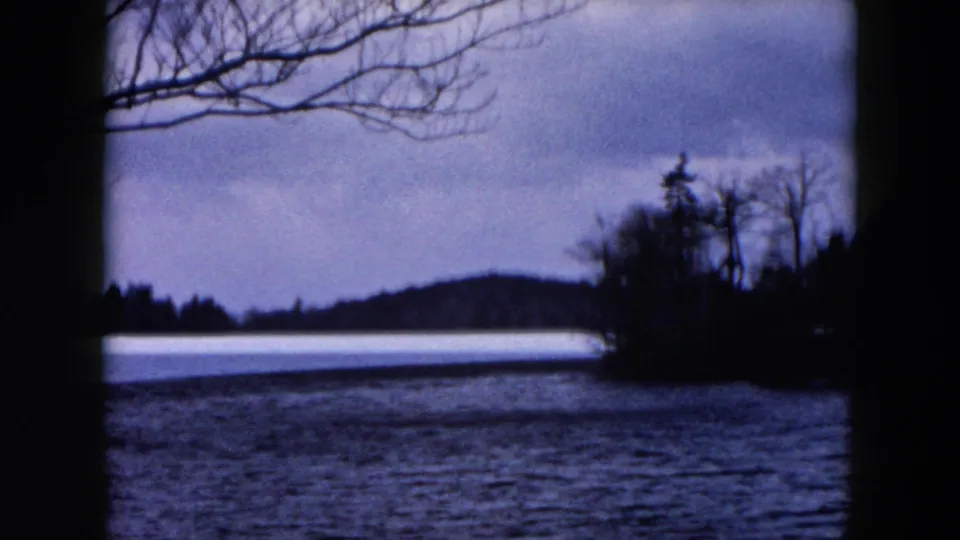 1957 8mm frame from the Hicksville, New York reel showing a lake, bare winter branches, distant wooded shore, and muted sky.