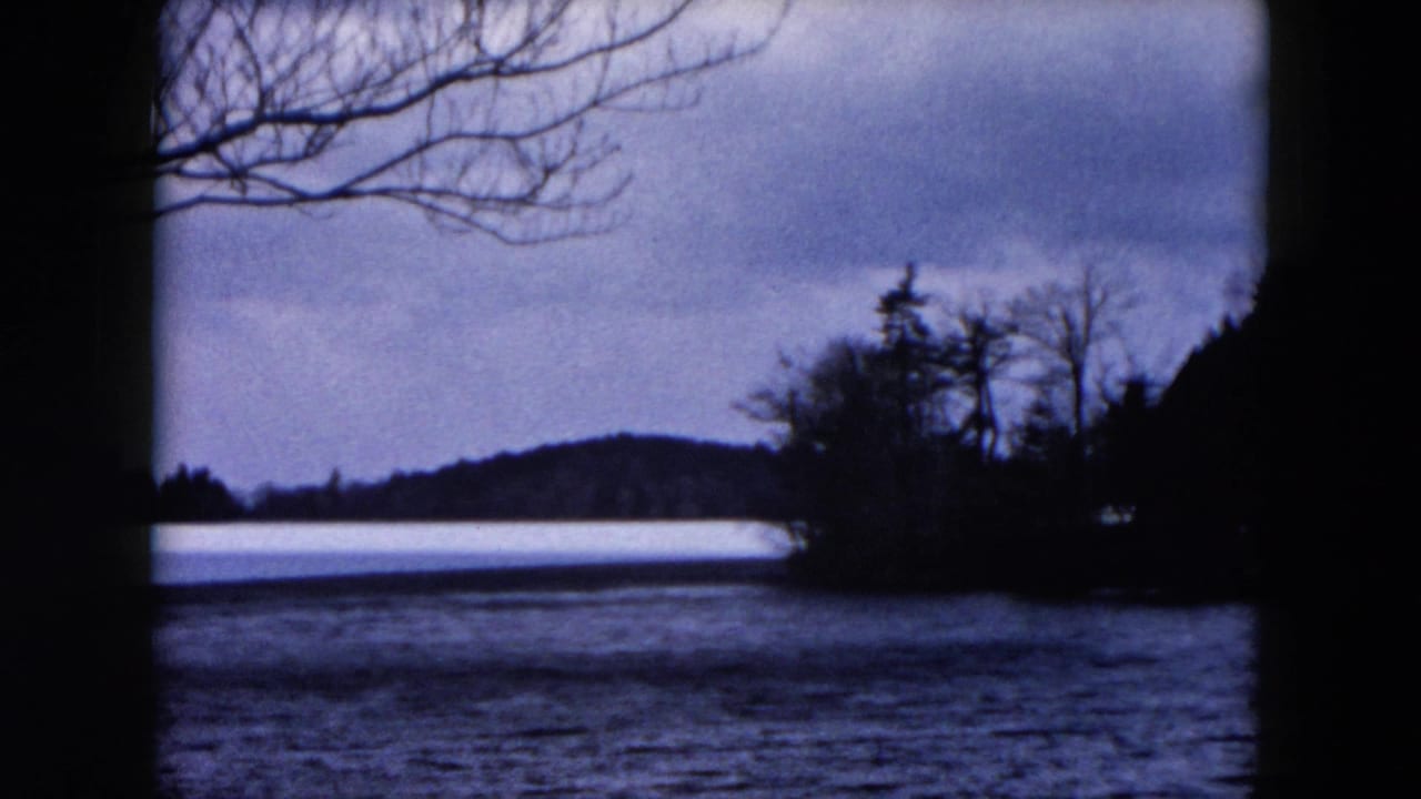 1957 8mm frame showing lake water, a distant tree-lined shore, bare branches, and heavy dark foreground framing.