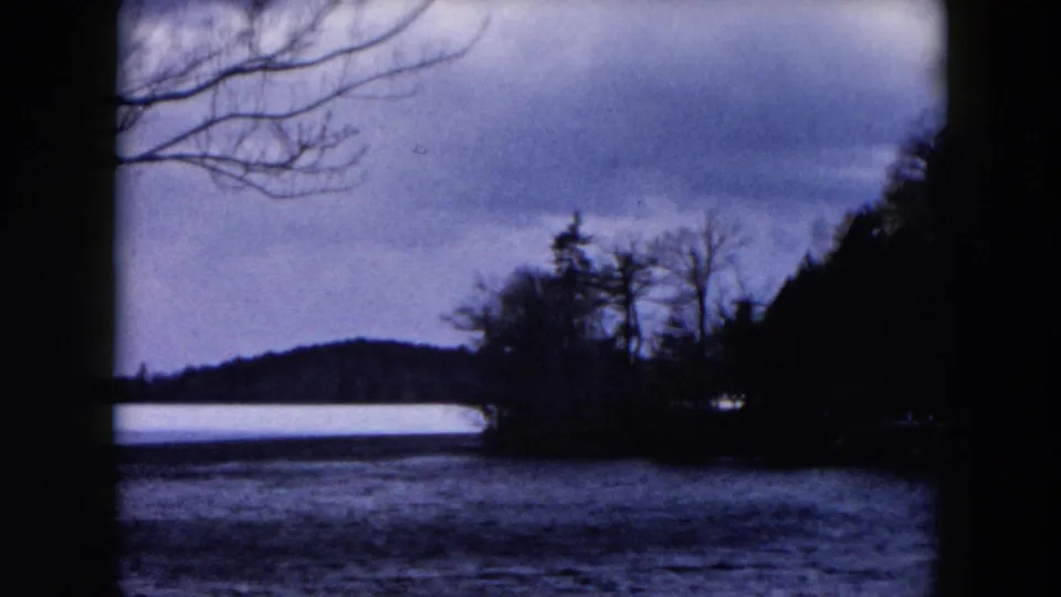 1957 Hicksville-area home-movie frame with a muted lake view, bare branches, distant wooded rise, and overcast sky.