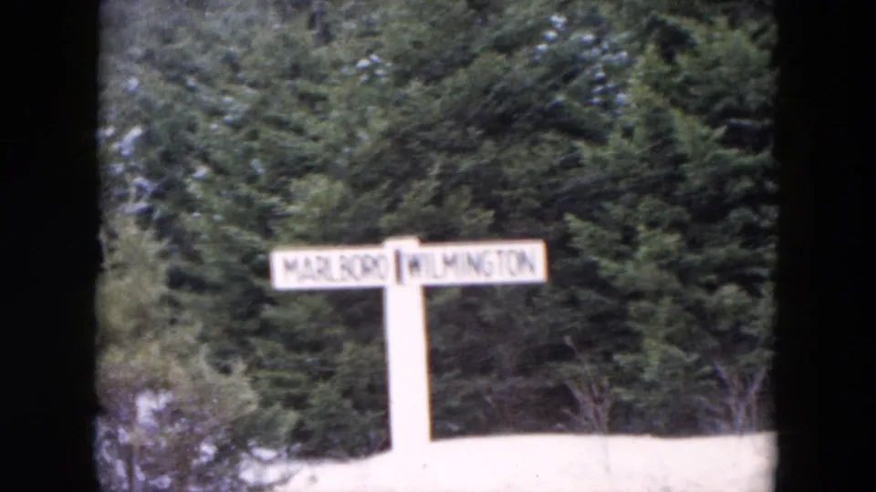 1957 8mm frame from the New York home-movie reel showing a roadside Marlboro Wilmington sign against evergreens and snow.