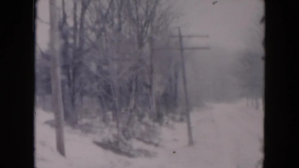 The frame shows a snow-covered roadside with leafless trees, utility poles, and a hazy road surface in archival 8mm color.