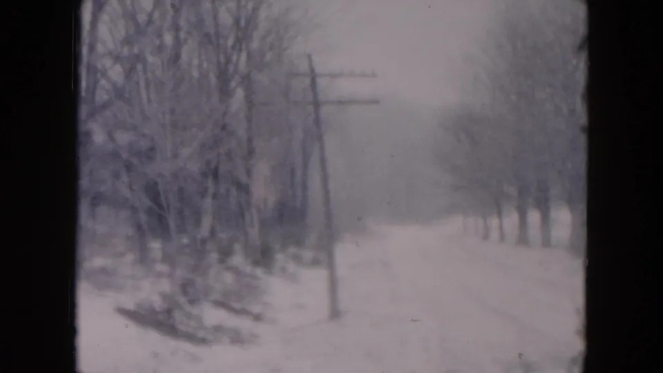 A quiet winter road recedes through snow, with bare trees, utility poles, and muted gray light in a 1957 home-movie frame.
