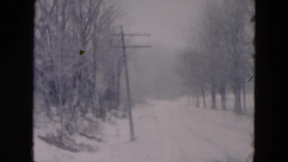 1957 8mm home-movie frame from metadata showing a snow-covered road in Hicksville, New York, with bare trees and a foreground utility pole.