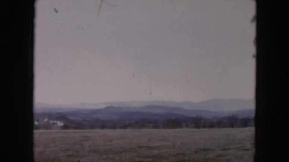 Muted 8mm home-movie frame of an open New York landscape, with fields and a distant ridgeline under a gray sky.