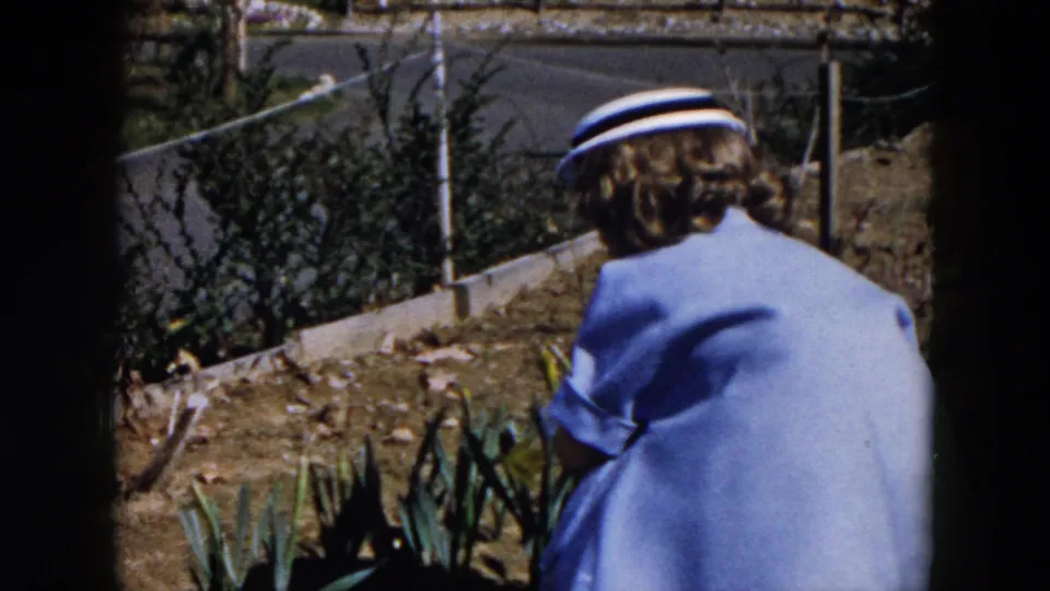 Seen from behind, the woman in a pale blue dress and white hat works close to a planted bed in the Hicksville backyard.