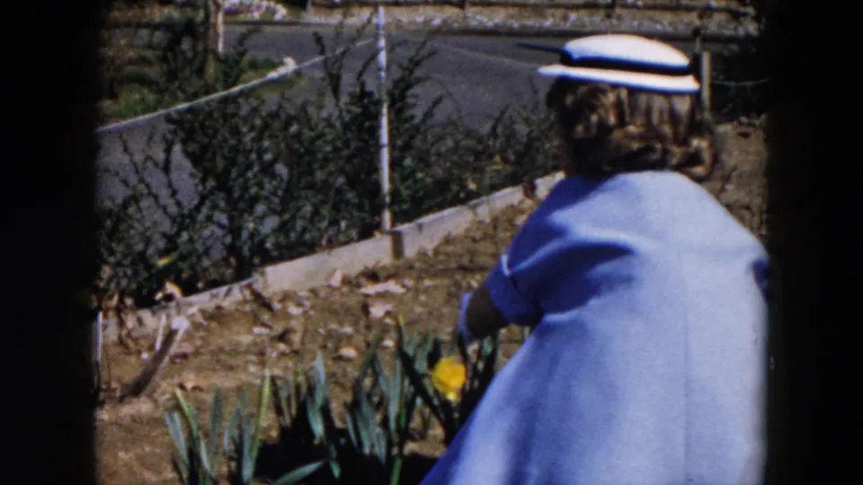 The woman reaches into the garden bed while wearing a pale blue dress, white hat, and gloves, with a chain-link fence behind her.