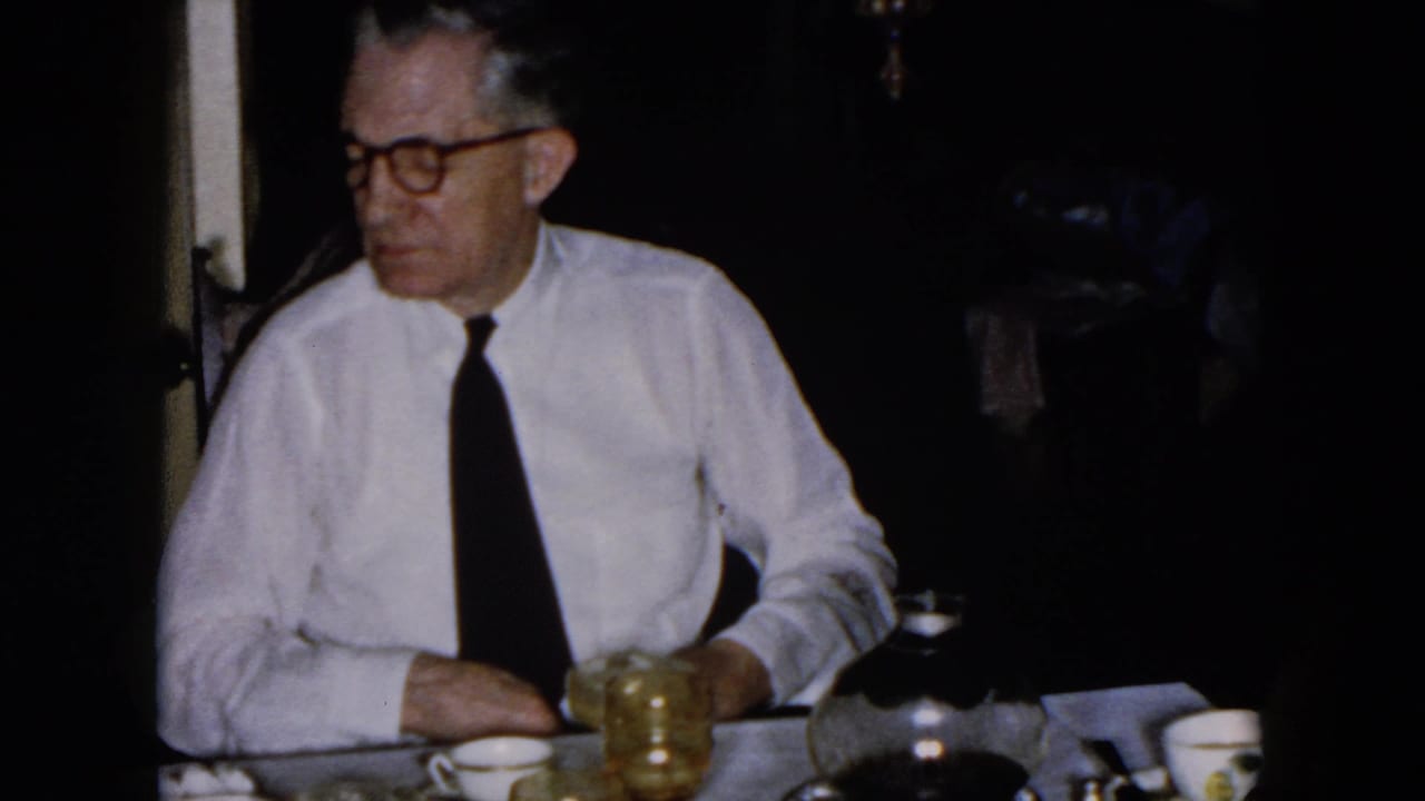 Older man in glasses sits at a dim dinner table with a drink.