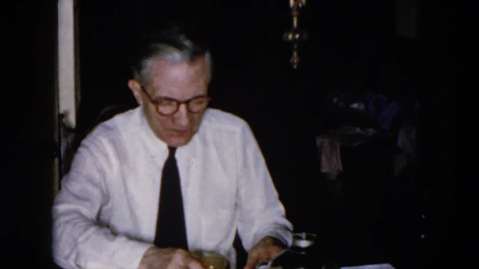 The man sits at the table in the Hicksville interior, looking down toward a glass and table items under low light.