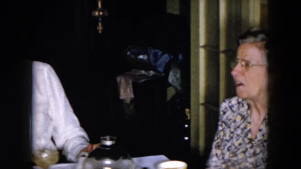A woman in glasses and a patterned blouse sits near the table in the 1957 Hicksville home-movie interior, partly obscured by shadow.