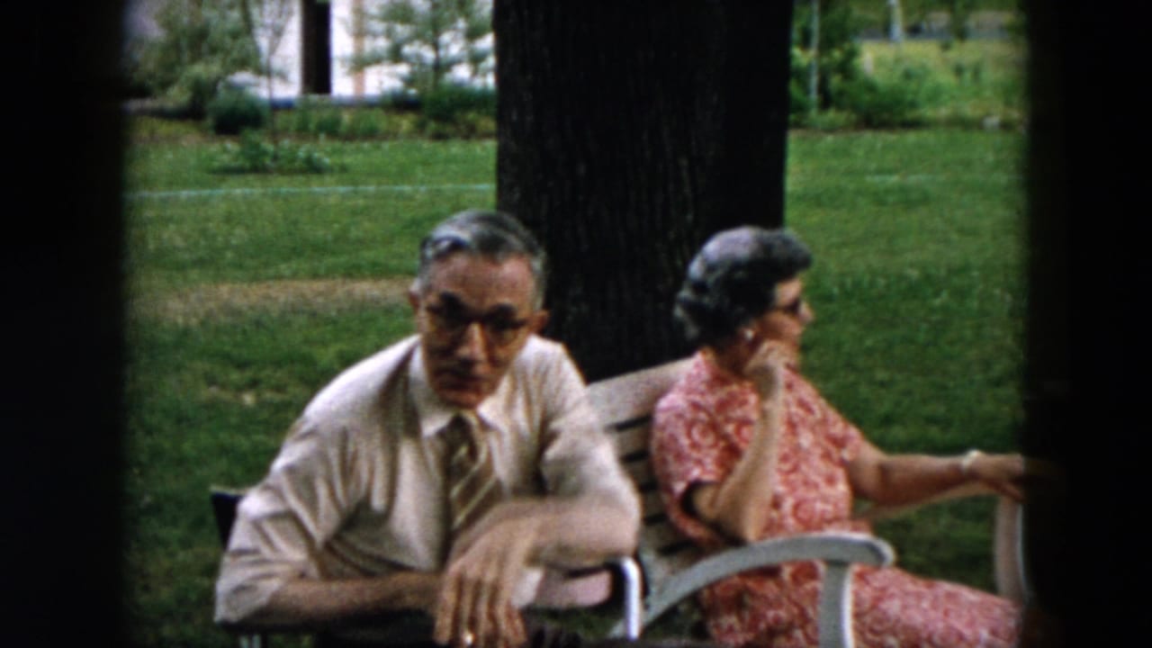 Older man in shirt and tie sits beside an older woman in a red patterned dress outdoors.