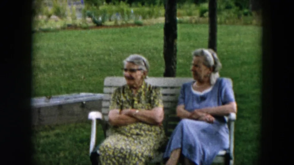 Two older women in patterned and blue dresses sit outdoors in Hicksville, New York, amid grass and trees.