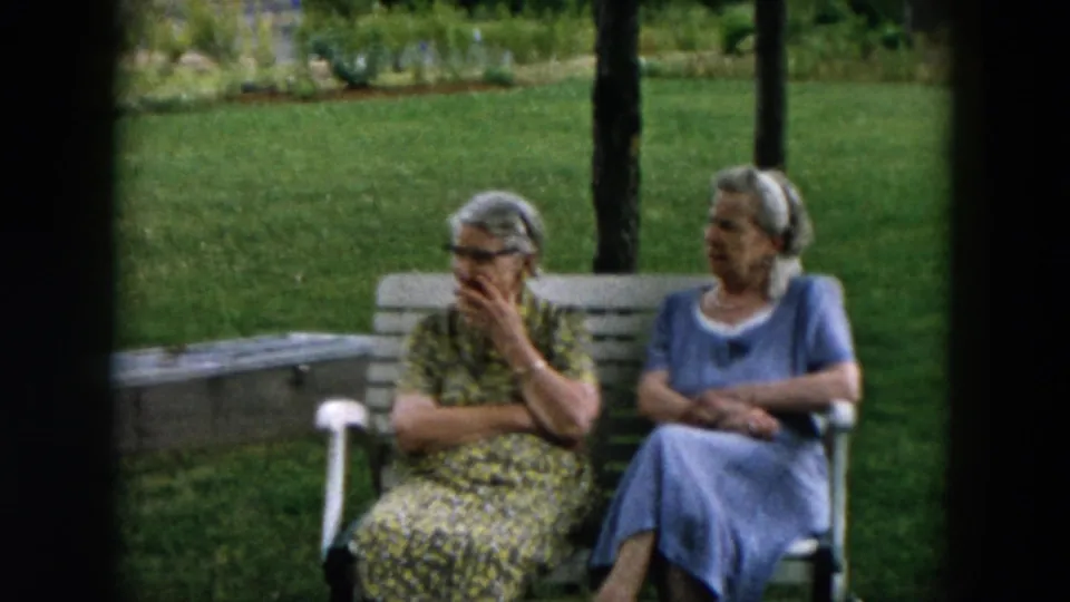 Two older women seated in a green outdoor setting, one gesturing near her face during conversation.