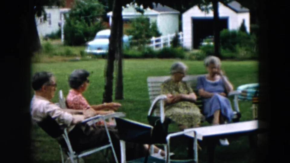 Adults gather in a Hicksville backyard in 1957, seated on lawn chairs and a bench with grass, houses, trees, and a white fence behind them.