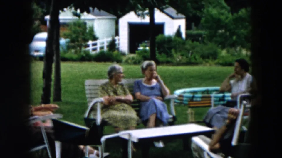 The 1957 Hicksville home-movie frame shows adults sitting in a grassy yard near a striped inflatable pool, with houses and a white fence in view.