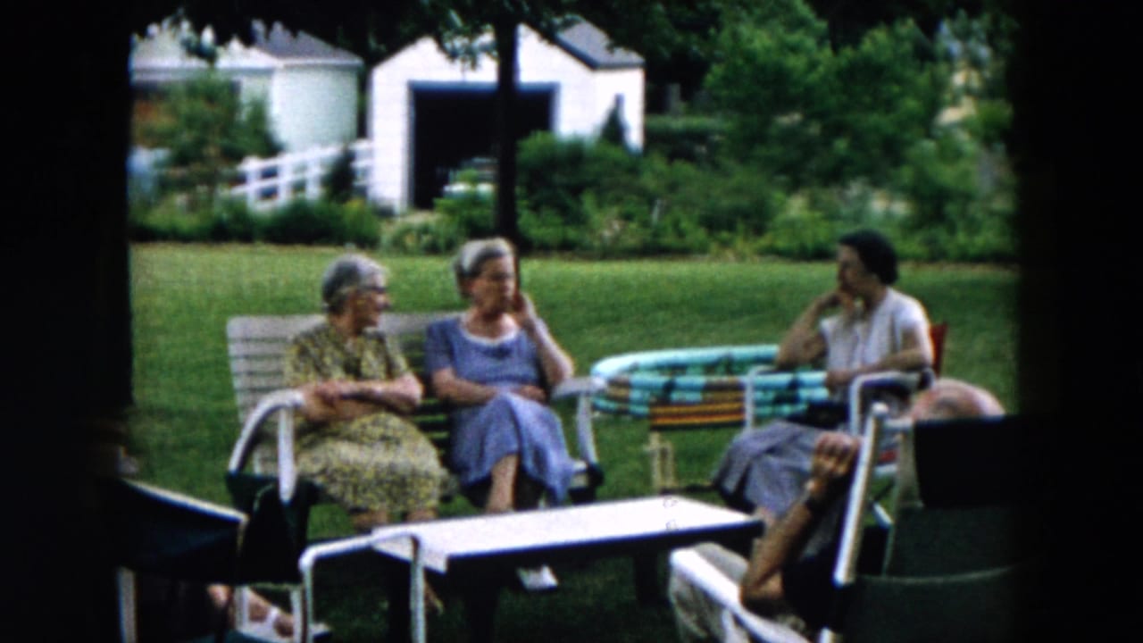 Adults sit outdoors in lawn chairs and on a bench beside a striped table.