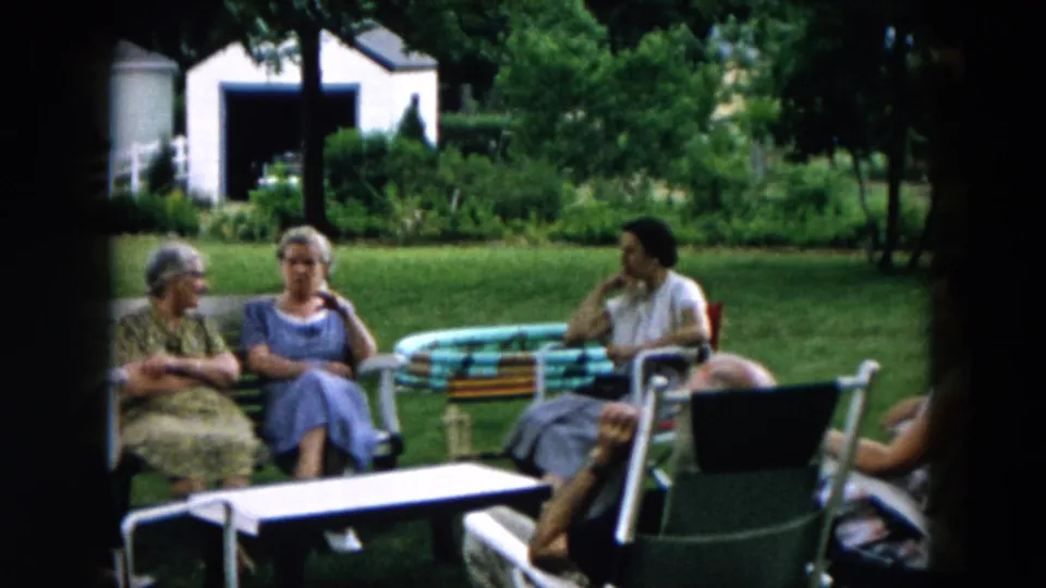 A group of adults relaxes in a 1957 Hicksville backyard, with lawn chairs, a striped table, trees, and a white shed visible.