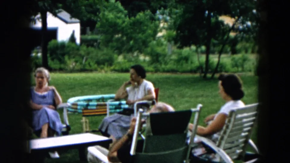 Adults are seated in a 1957 Hicksville backyard, framed from a home-movie viewpoint with lawn furniture and greenery visible.