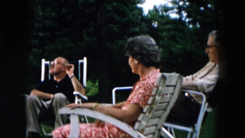 Adults sit in profile in a Hicksville backyard in 1957, with white lawn chairs, trees, and shaded grass behind them.