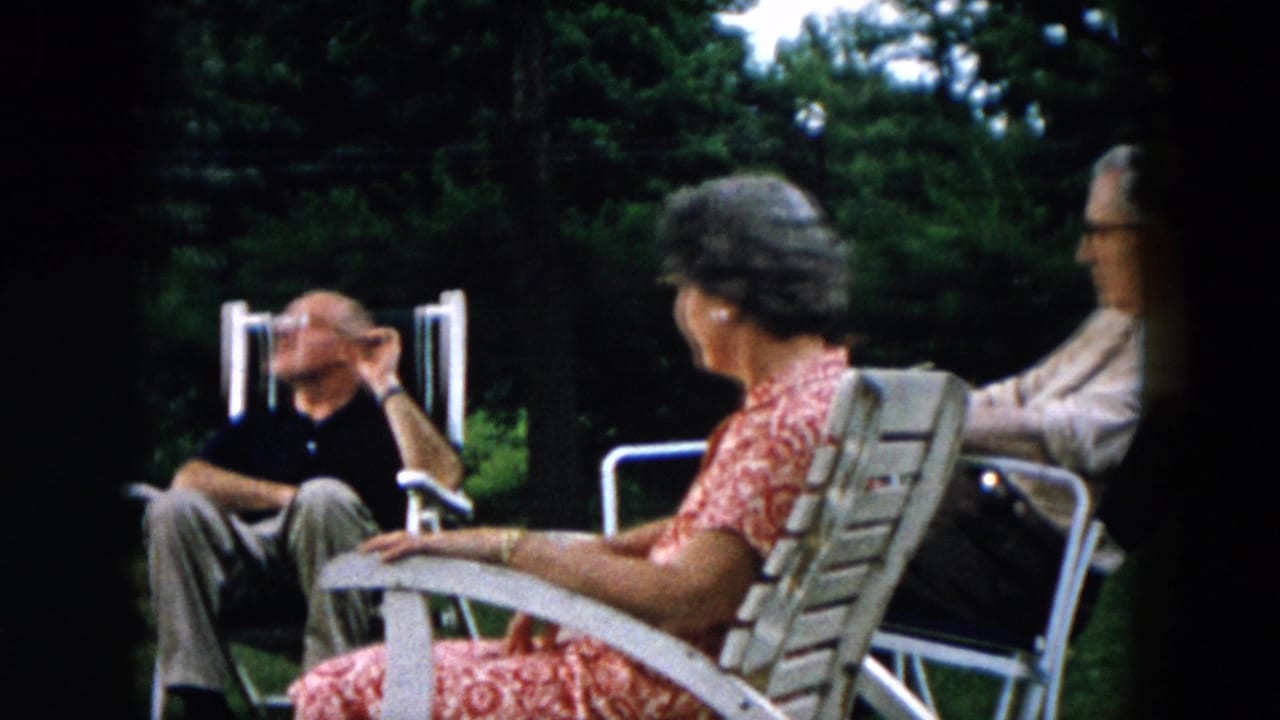 Adults seated in white lawn chairs in a backyard, with one man gesturing while others listen.
