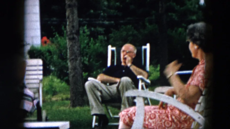 A 1957 Hicksville backyard view shows a seated man in a dark shirt and a woman in a red patterned dress gesturing beside lawn chairs.
