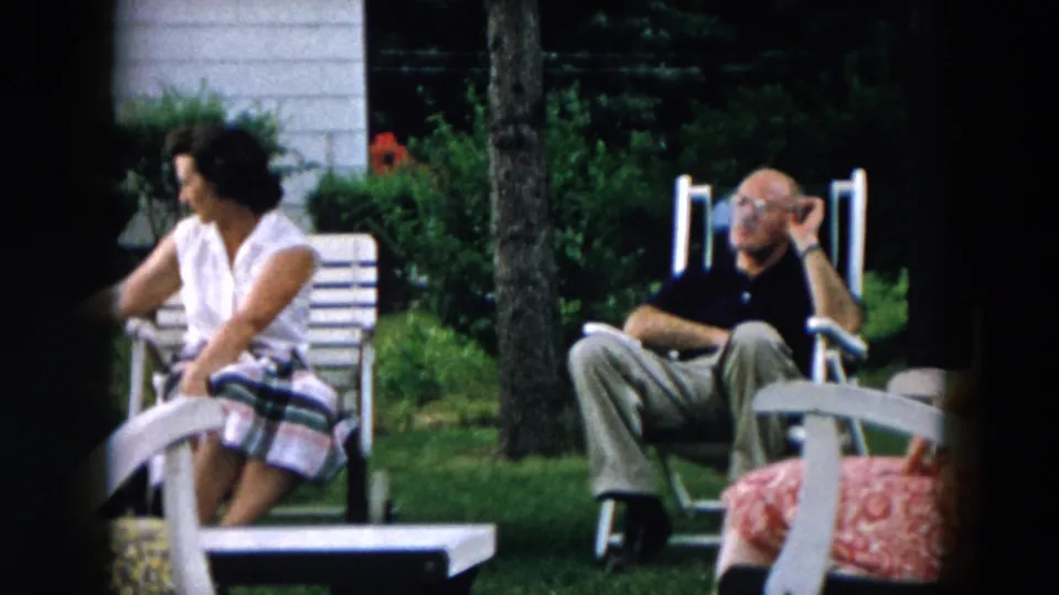 In this 1957 Hicksville home-movie frame, a woman in a white top and plaid skirt sits near a man in a dark shirt on the lawn.