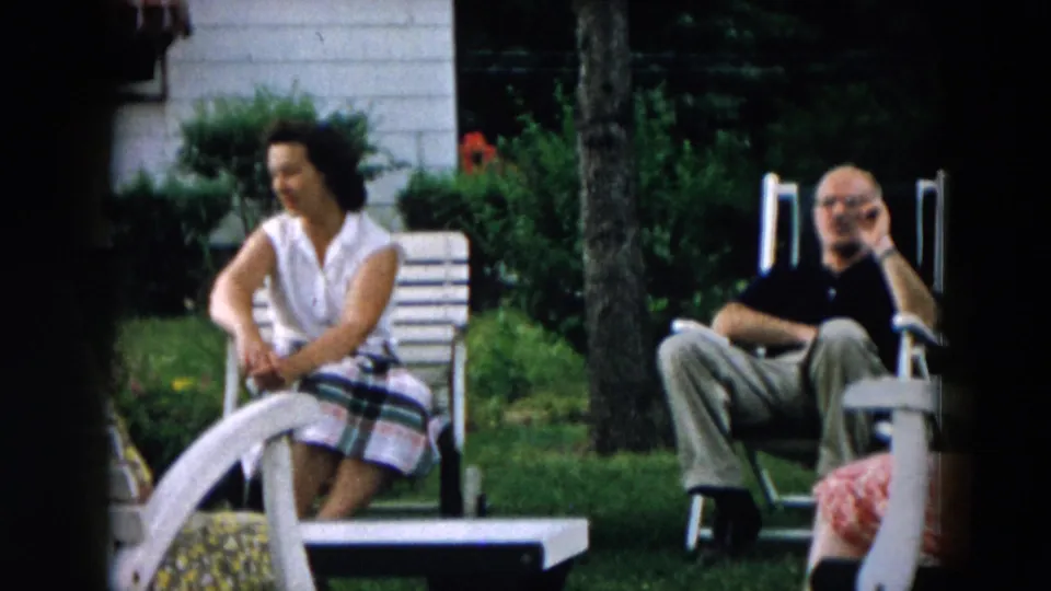 A 1957 Hicksville backyard frame shows a woman seated with hands together near a man in a white lawn chair and another adult partly visible.