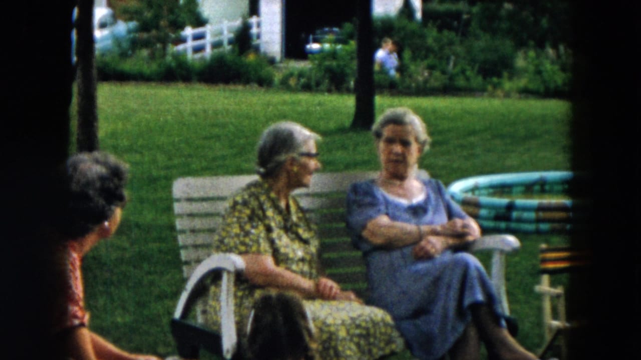 Three elderly women sit and talk near a white bench on a green lawn.