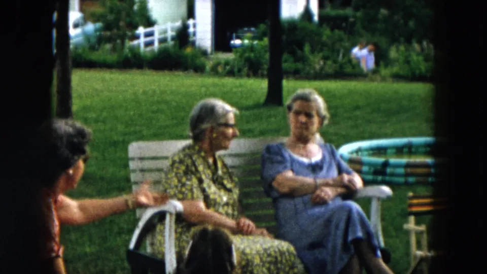 A woman in a red patterned dress gestures while two elderly women sit on a white bench in a 1957 Hicksville backyard.